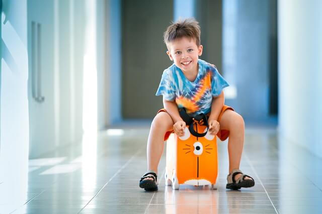 Kind sitzt auf einem rollbaren Koffer und fährt mit diesem durch einen Gang